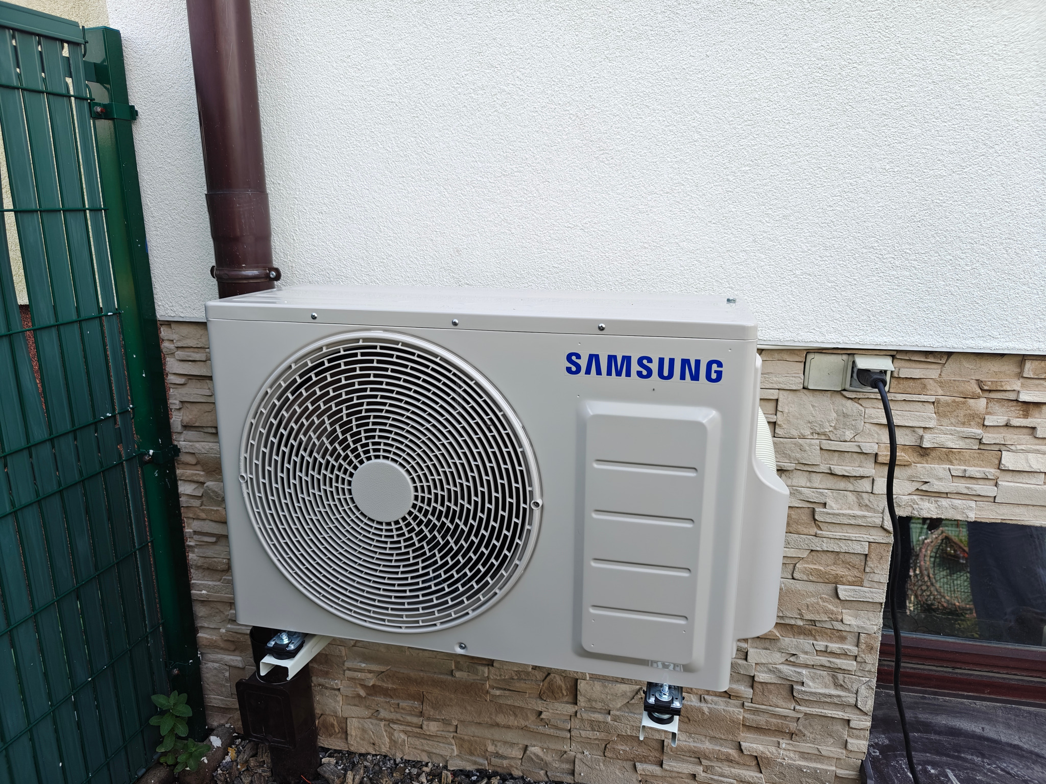 Zewnętrzna jednostka klimatyzacji Samsung zamontowana na ścianie z elementami dekoracyjnymi z kamienia, obok rynny i zielonego ogrodzenia.