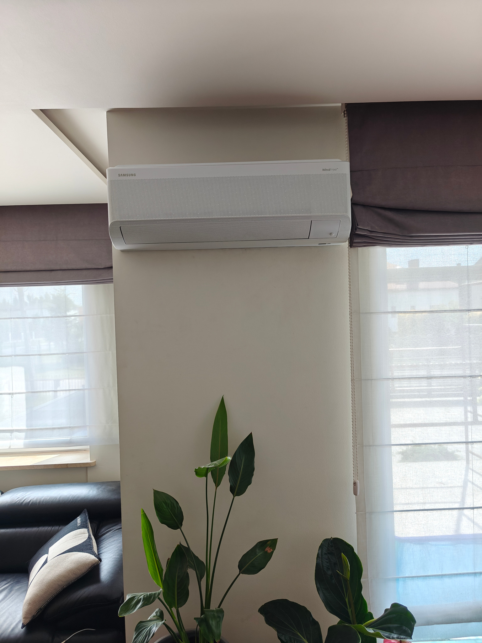 Biała klimatyzacja Samsung WindFree na ścianie w salonie z roletami rzymskimi i rośliną doniczkową. Widok z dołu na urządzenie.