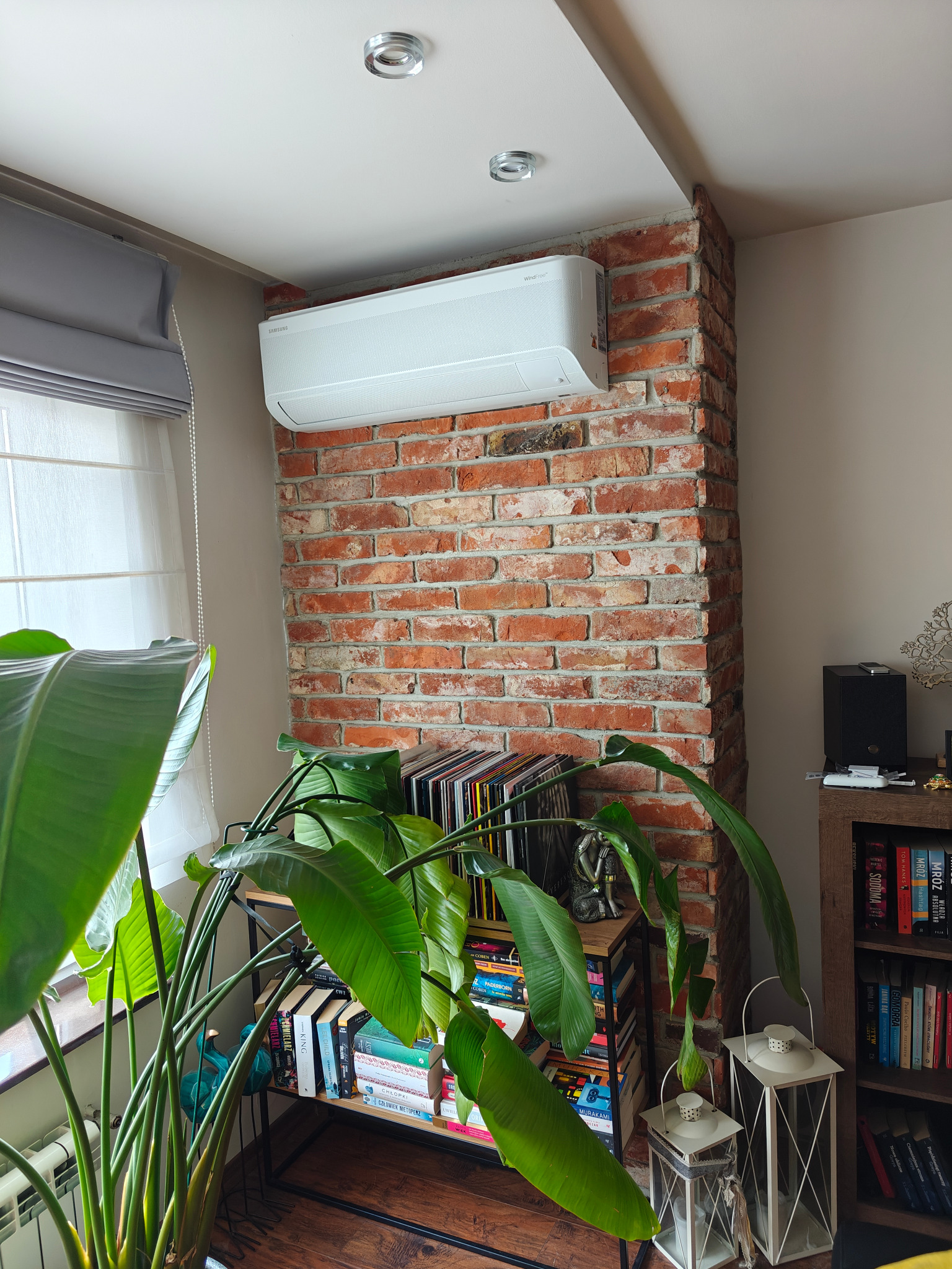 Biała klimatyzacja Samsung WindFree zamontowana na ceglanej ścianie w pokoju z rośliną, książkami i lampami. Widok na okno z roletami.