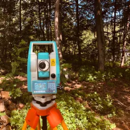 Teodolit elektroniczny ustawiony na pomarańczowym statywie w zalesionym terenie, z widocznym ekranem i klawiaturą, a w tle drzewa i runo leśne.