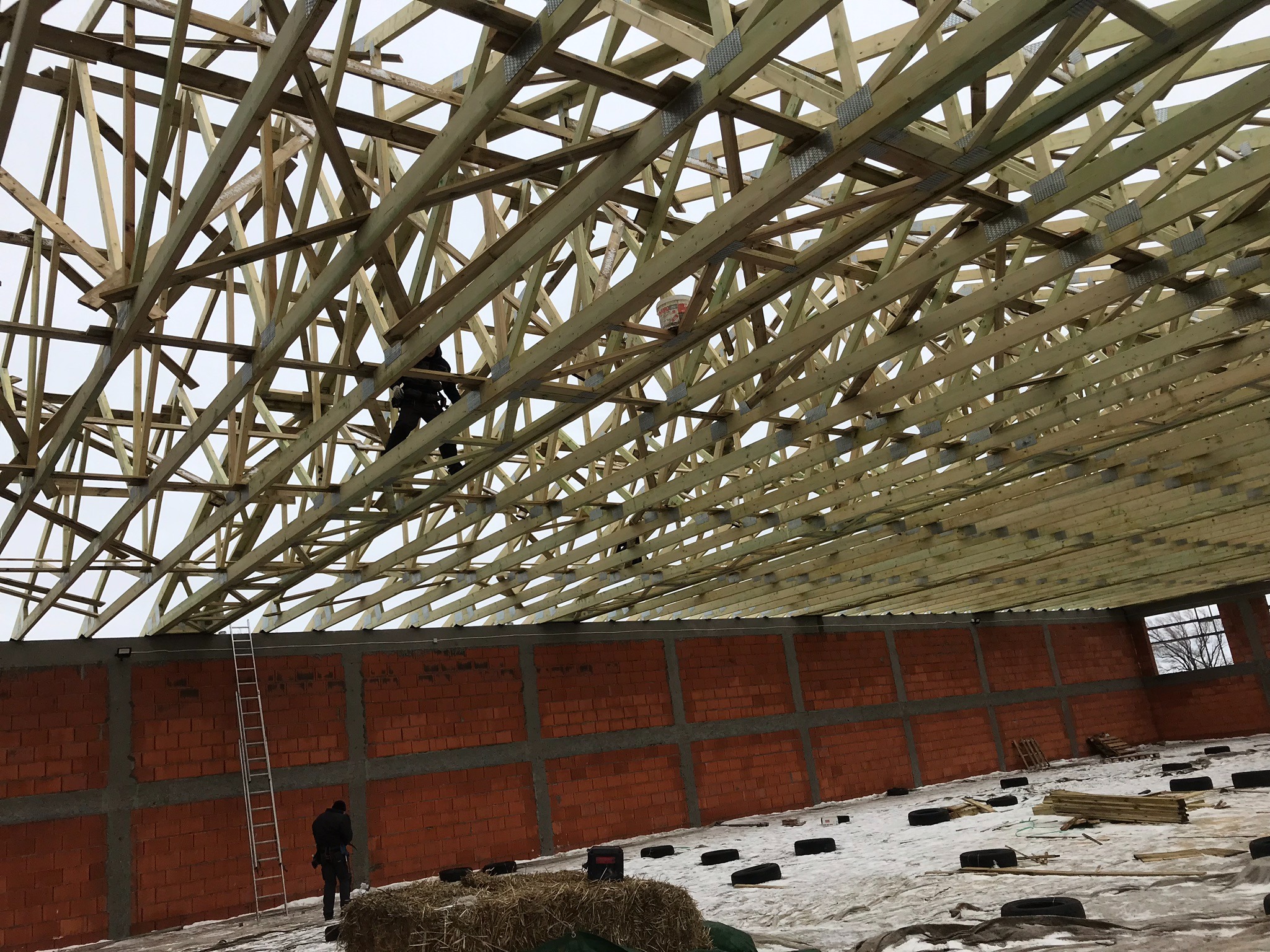 Konstrukcja drewniana dachu w trakcie montażu, widoczny pracownik na wysokości, mur z czerwonej cegły i częściowo ośnieżony teren budowy.