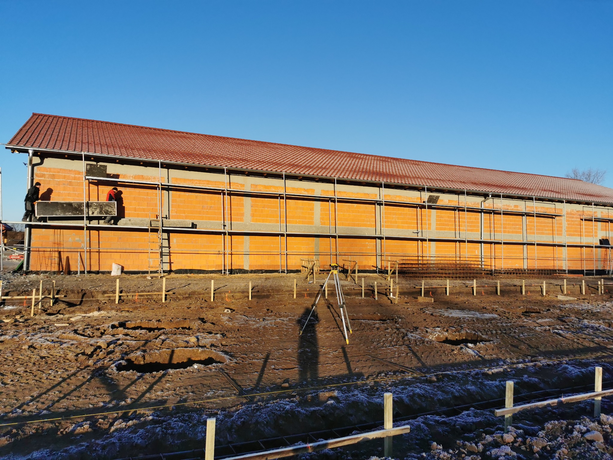 Budowa domu jednorodzinnego z czerwonej cegły, widoczne rusztowania, dwóch pracowników na rusztowaniu i niwelator na ziemi.