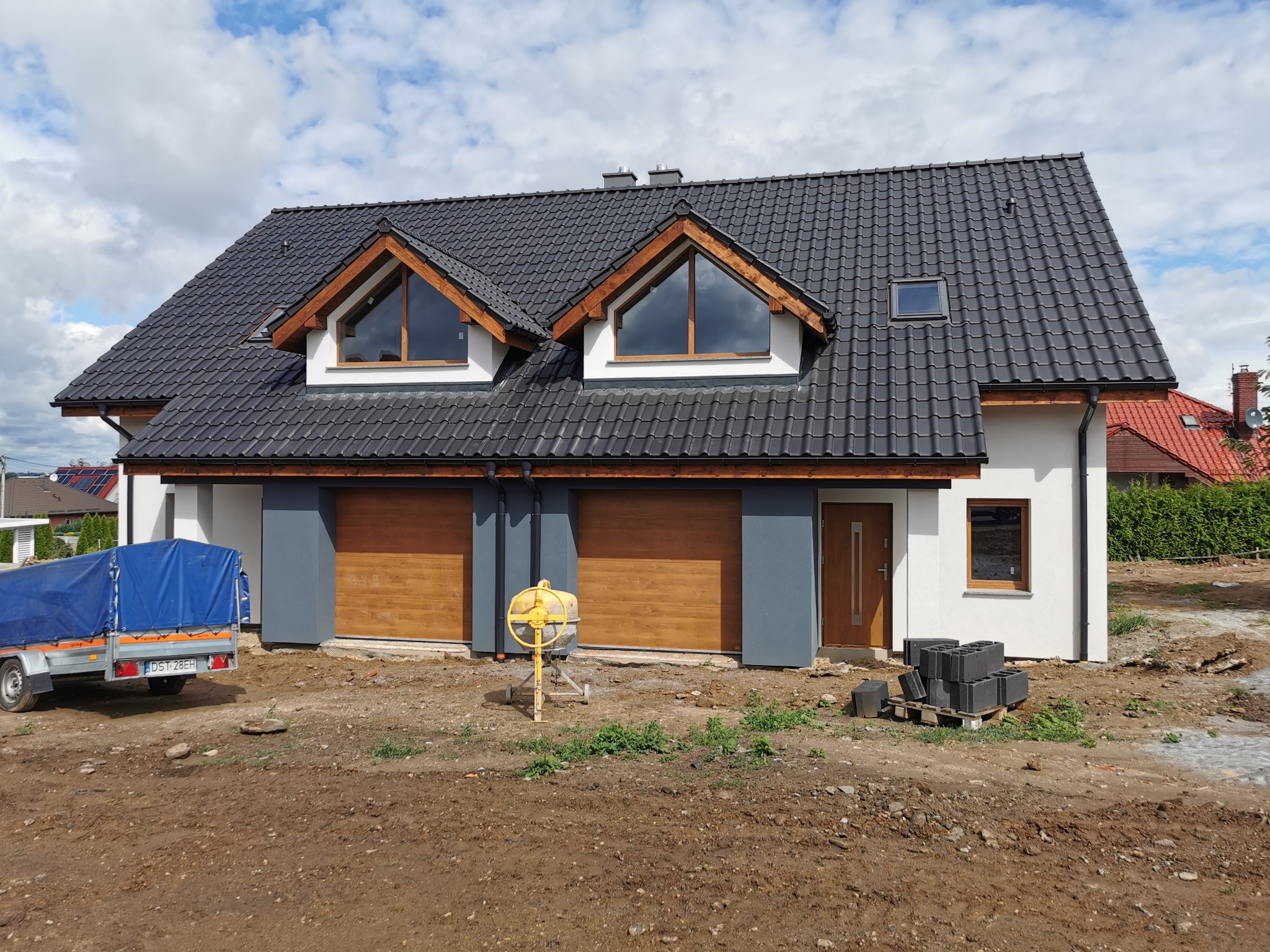 Nowoczesny dom jednorodzinny z ciemną dachówką i drewnianymi akcentami, widoczny na etapie prac wykończeniowych z betoniarką i przyczepą na placu budowy.