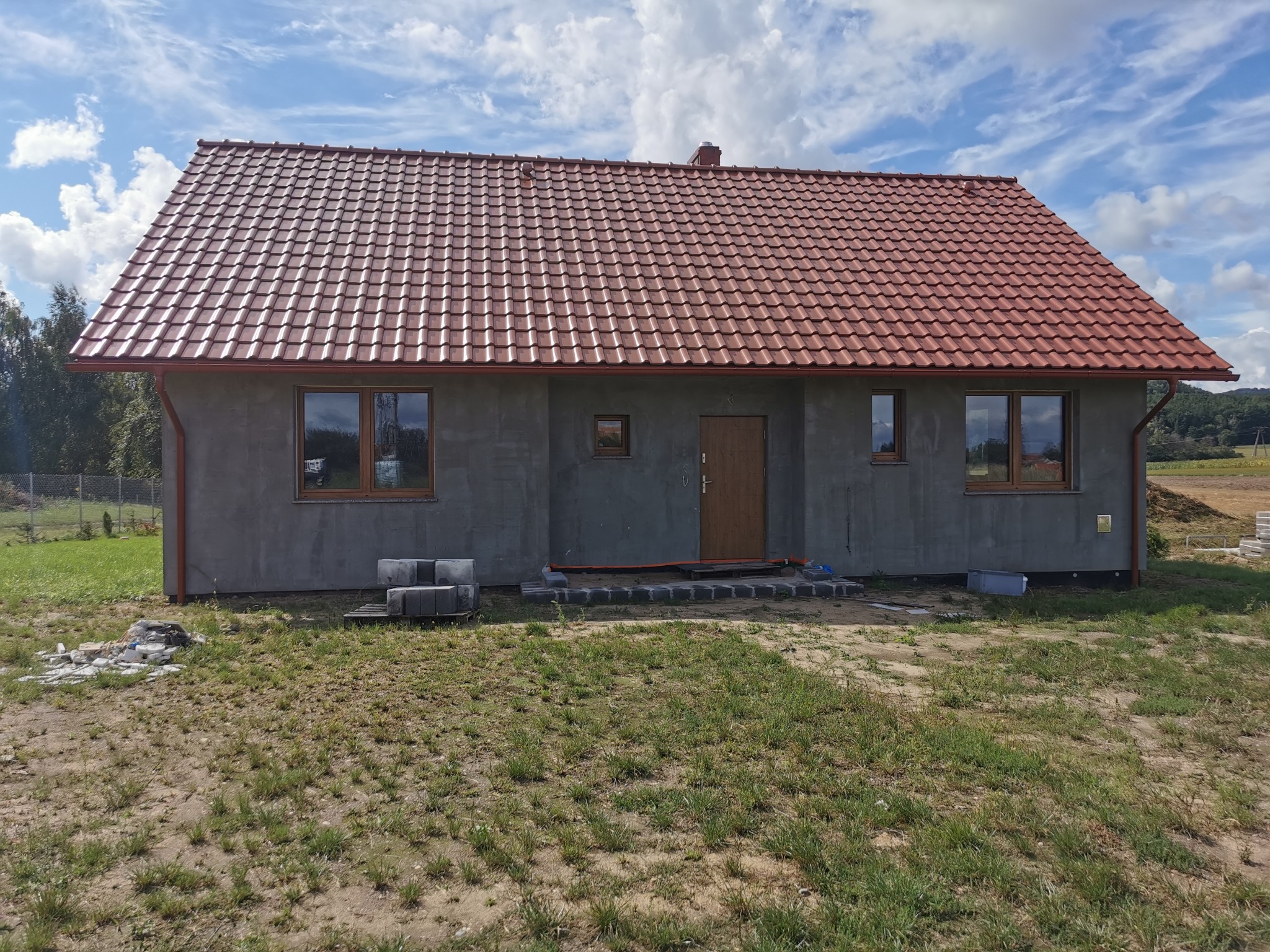 Surowy dom jednorodzinny z czerwoną dachówką, widoczne okna z drewnianą stolarką, częściowo wykończone wejście z kostki brukowej, w tle błękitne niebo z chmurami.