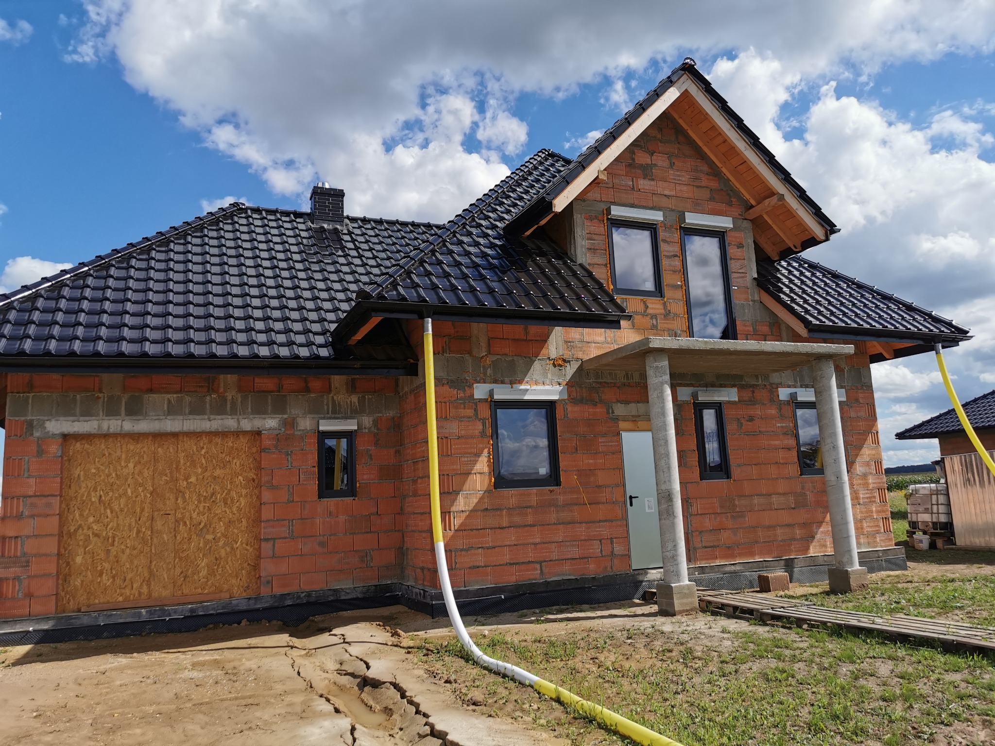 Surowy budynek jednorodzinny z czerwonej cegły, z czarną dachówką, częściowo otynkowany, z żółtym wężem na trawniku.