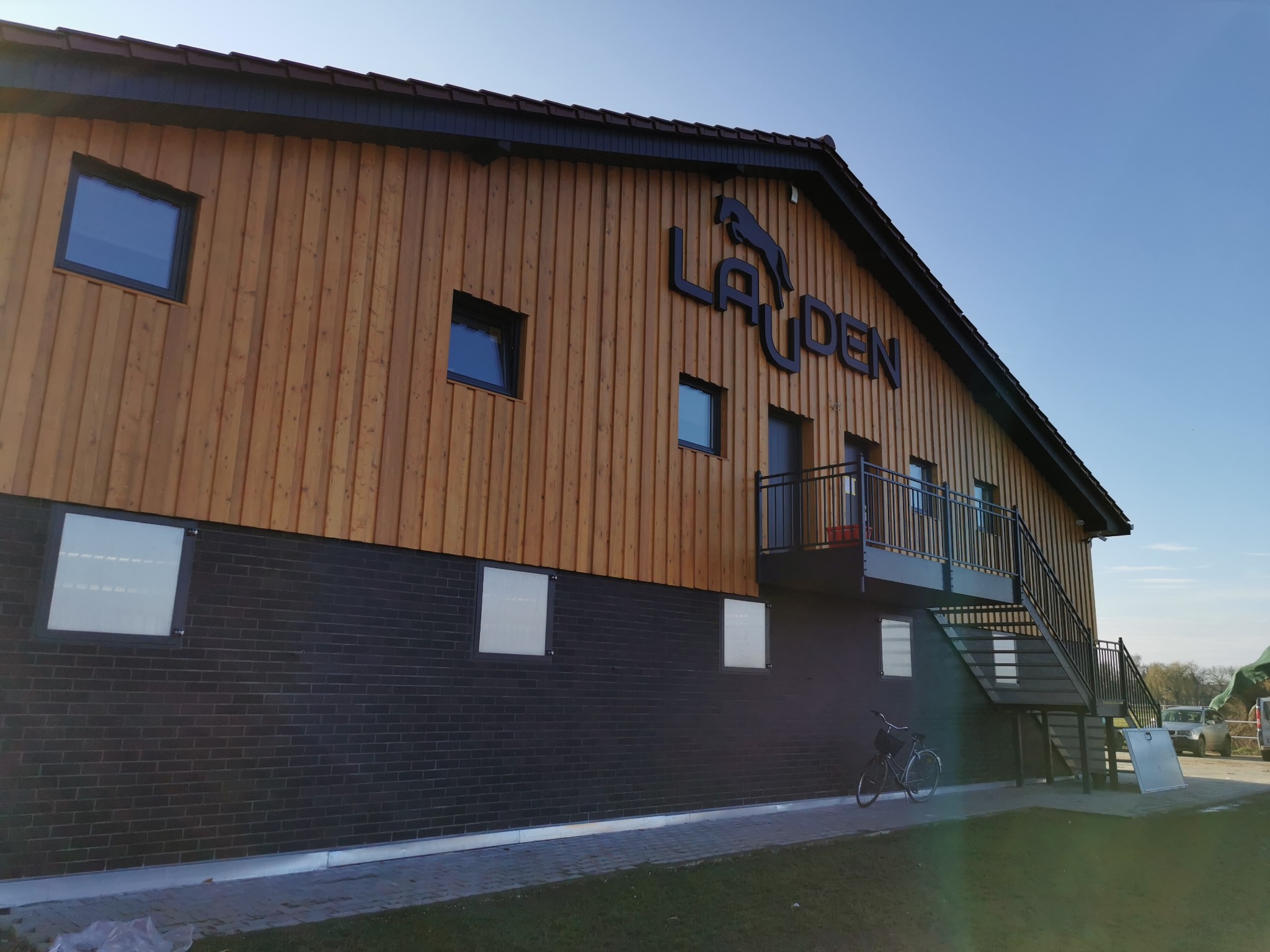 Nowo wybudowany budynek z drewnianą elewacją w Przewornie, z czarnym napisem 'LAUDEN' i symbolem konia, balkonem i zewnętrznymi schodami, stojący na tle błękitnego nieba.