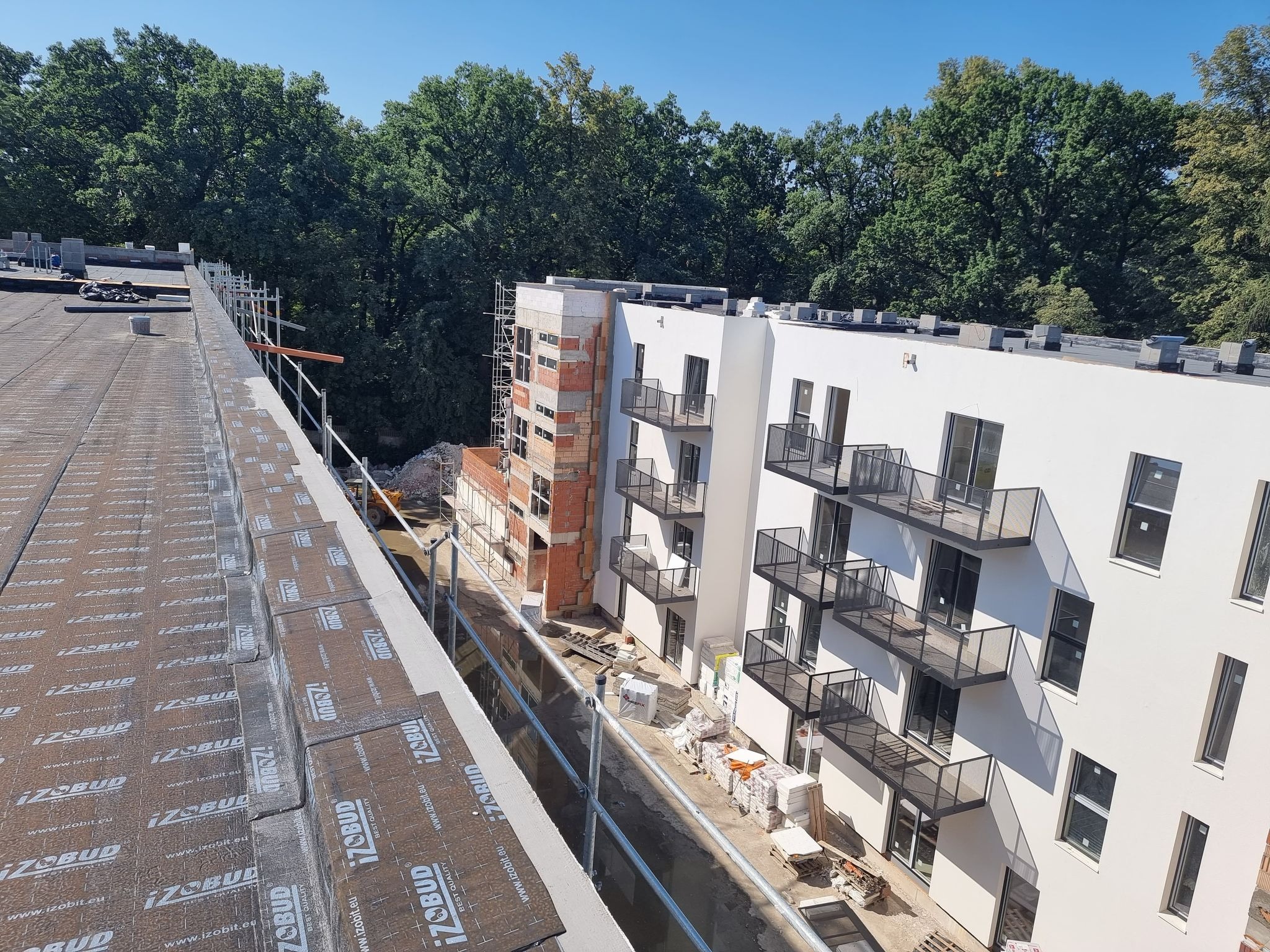 Montaż izolacji na dachu budynku wielorodzinnego w trakcie budowy, widoczne materiały izolacyjne IZOBUD, rusztowania i fragment elewacji z balkonami.