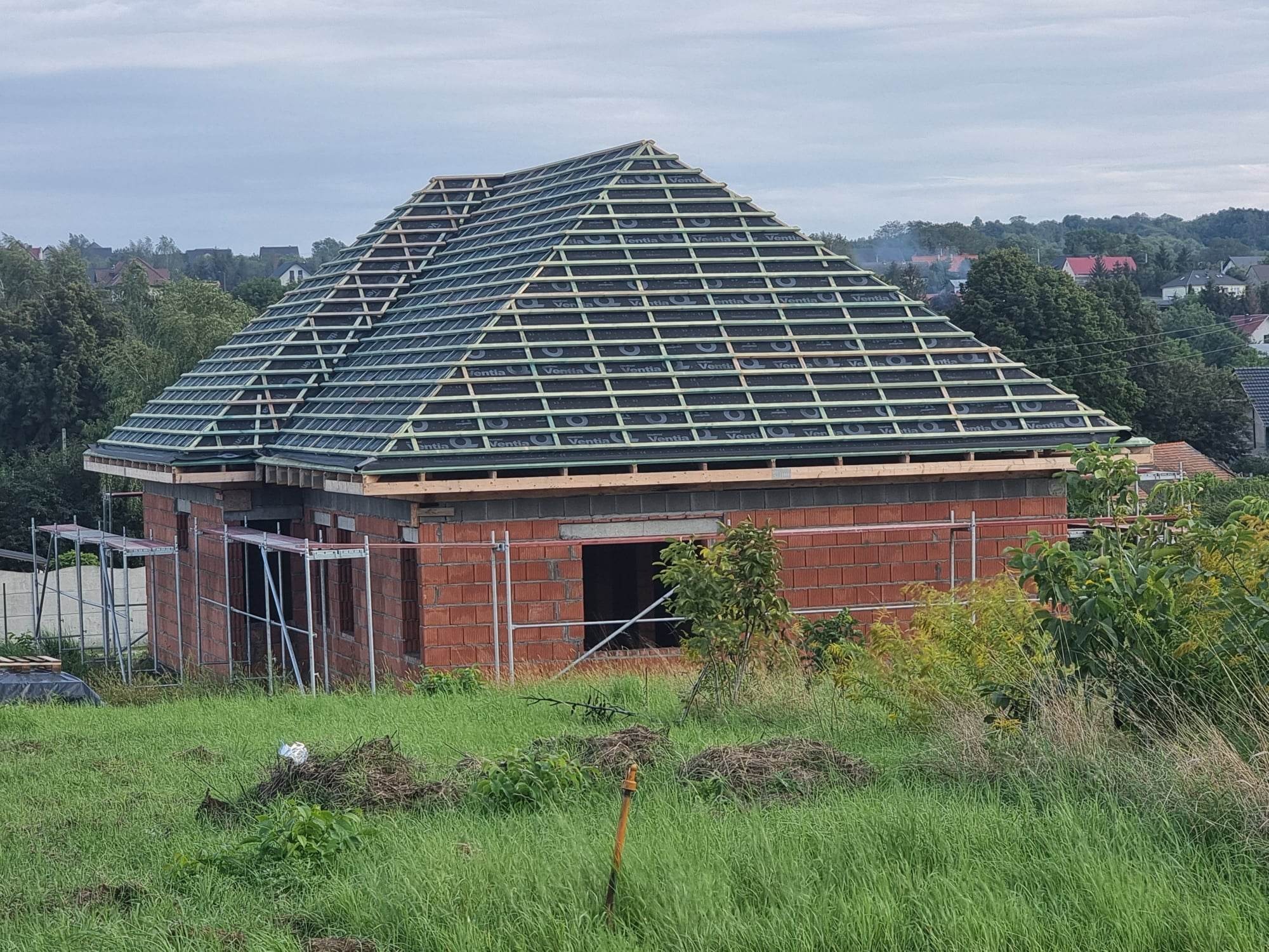 Surowa konstrukcja dachu z łatami i membraną na domu w trakcie budowy, widok z zewnątrz na tle zieleni.