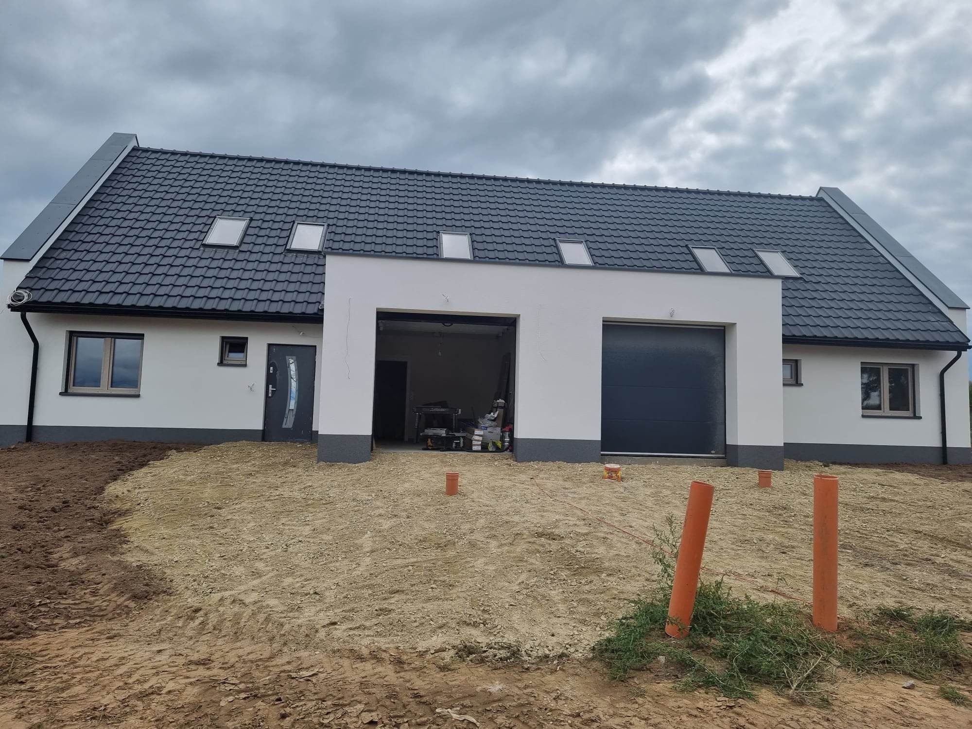 Nowoczesny dom jednorodzinny z ciemną dachówką i trzema oknami dachowymi, białą elewacją z szarym cokołem i dwustanowiskowym garażem w trakcie budowy; widoczne rury kanalizacyjne wystające z ziemi...