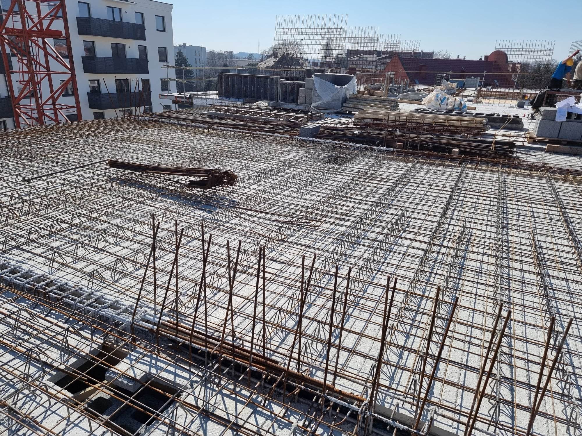 Zbrojenie stropu budynku wielorodzinnego w trakcie budowy, widok z góry na siatkę z prętów stalowych, w tle dźwig budowlany i budynki mieszkalne.