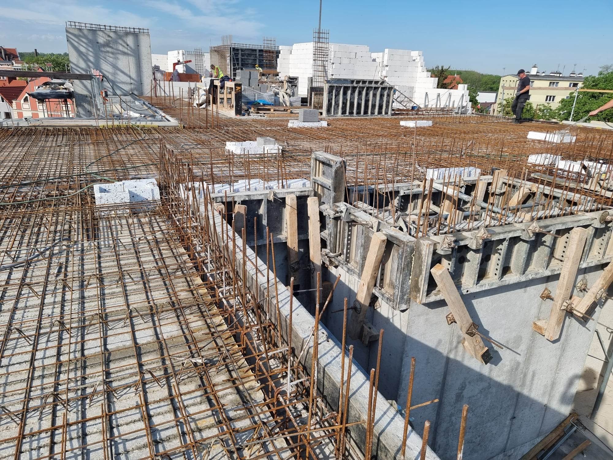 Zbrojenie stropu na budowie w trakcie betonowania, widoczne szalunki i siatka z prętów stalowych, w tle budynki mieszkalne i pracownicy.