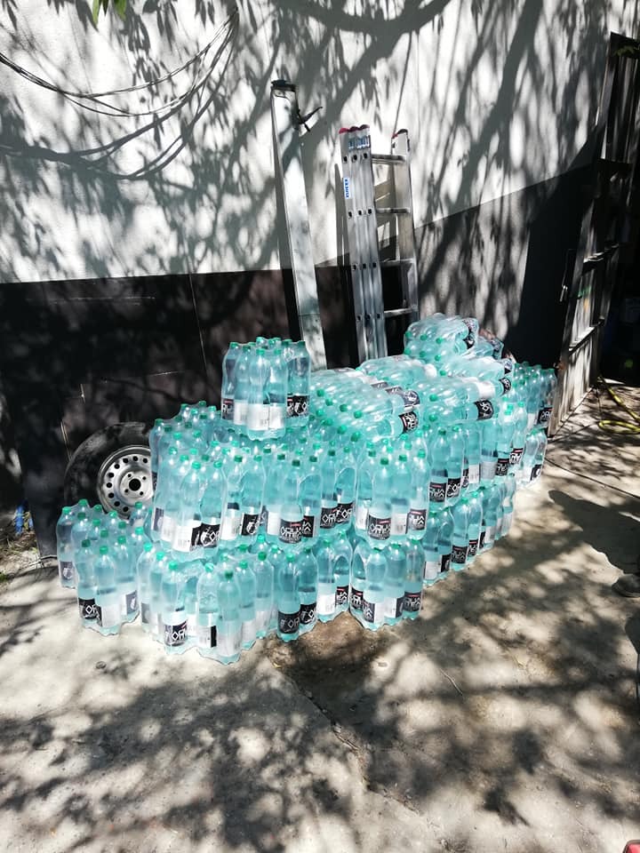 Stos butelek wody mineralnej, zapakowanych w folię, ułożony na ziemi w cieniu drzewa, z widoczną drabiną aluminiową w tle.