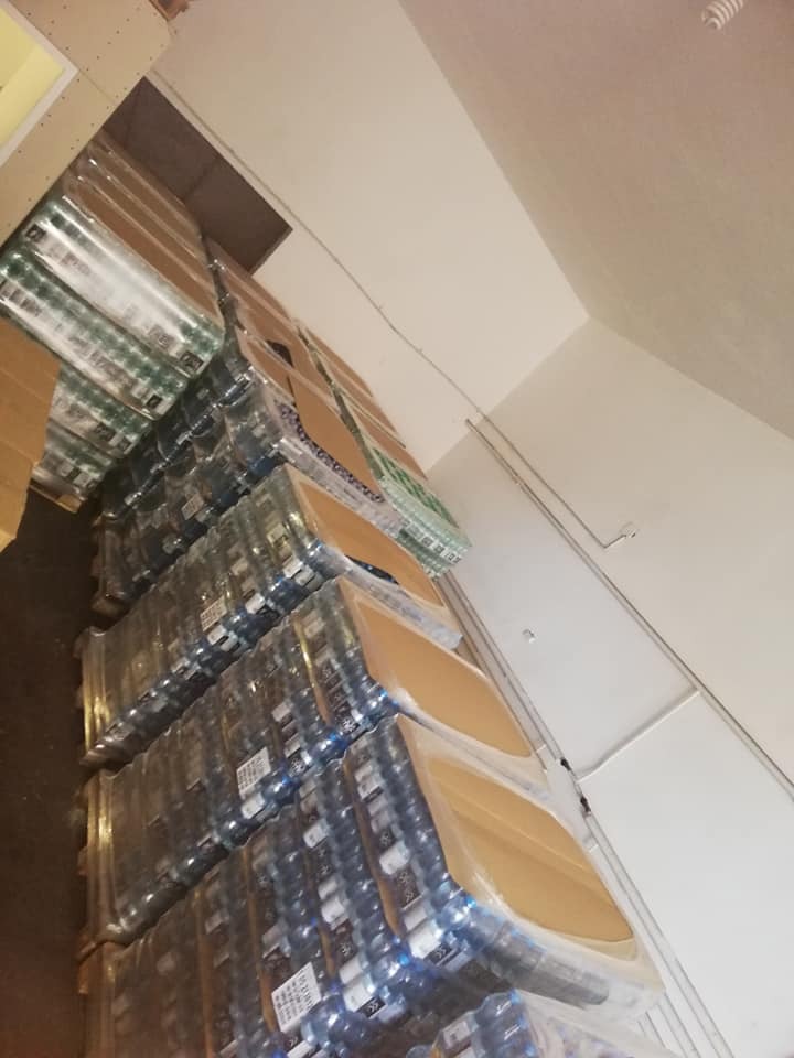 Stosy zafoliowanych palet z wodą butelkowaną w magazynie, widok z dołu, oświetlenie górne.