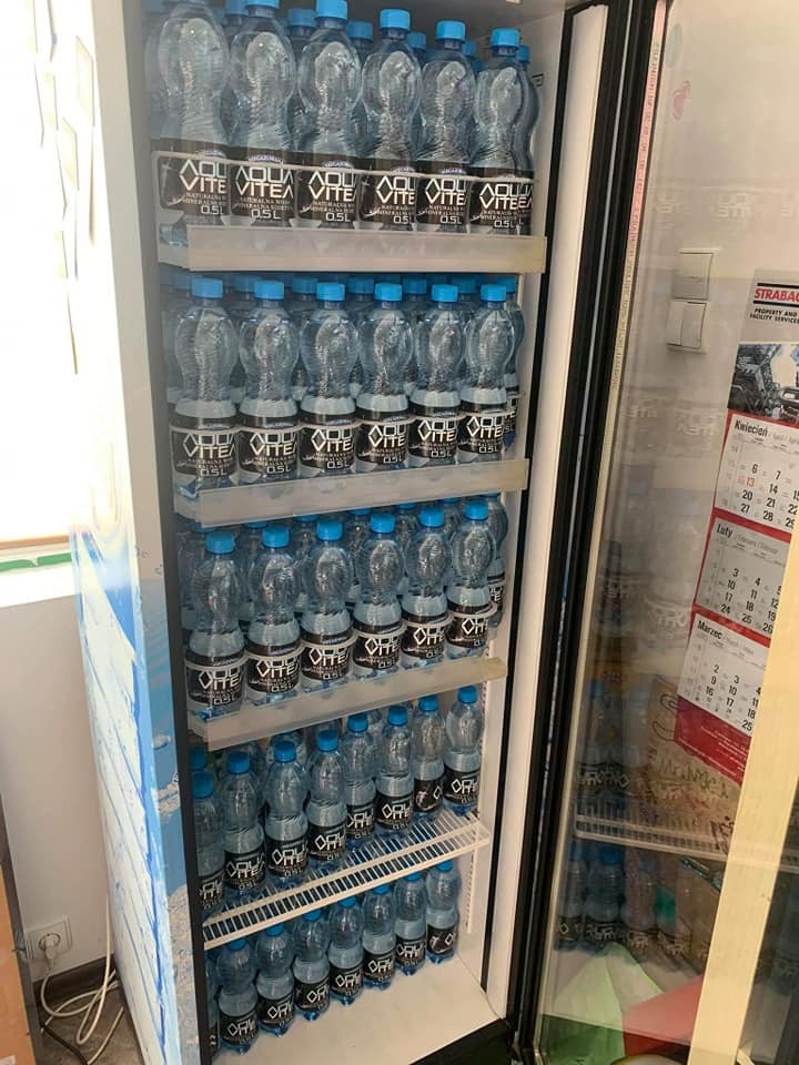 Chłodziarka wypełniona butelkami wody mineralnej Aqua Vitea 0.5L z niebieskimi zakrętkami, widoczny kalendarz za szybą chłodziarki.
