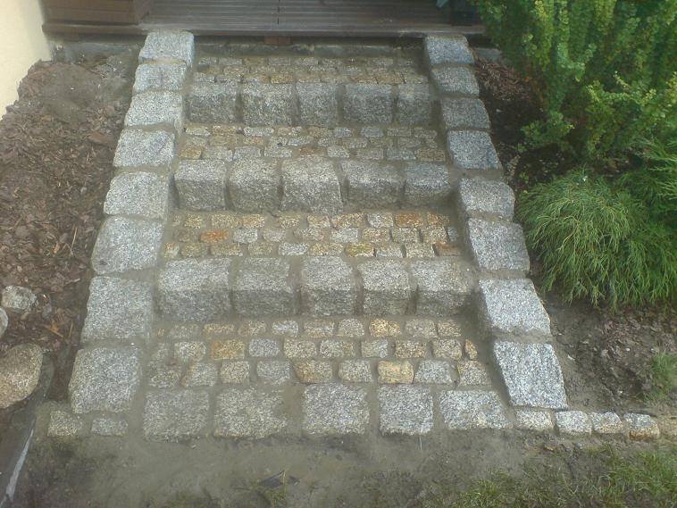 Schody wejściowe wyłożone kostką granitową w dwóch odcieniach szarości i beżu, z widocznymi spoinami i elementami zieleni w tle.