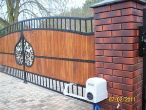 Brama wjazdowa z drewnianymi panelami i kutymi elementami dekoracyjnymi, zamontowana między dwoma murowanymi filarami z czerwonej cegły. Widoczny automat do otwierania bramy i fragment kostki...