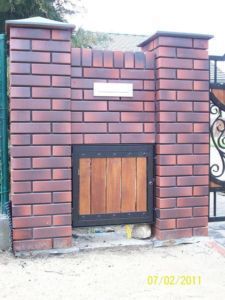 Fragment ogrodzenia z cegły klinkierowej z małą furtką z drewnianymi deskami w metalowej ramie. Nad furtką skrzynka na listy. Widoczne słupki ogrodzeniowe i fragment metalowej bramy w tle.