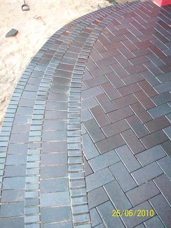 Ułożona, ciemnoszara kostka brukowa w wzór jodełki, z obrzeżem z prostokątnych kostek, widoczna łopata i piasek w tle.