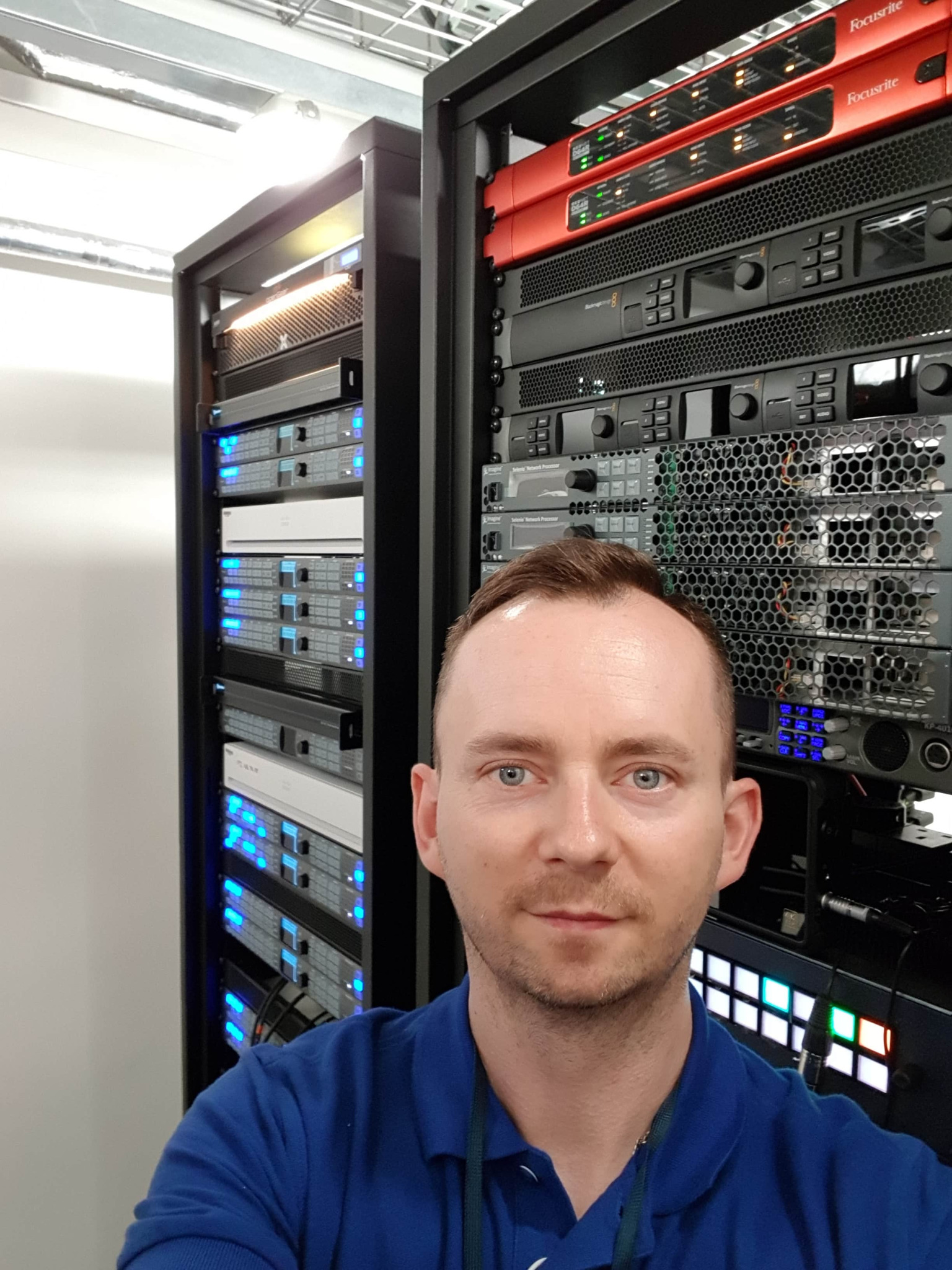 Mężczyzna robi selfie na tle szaf serwerowych z rozbudowaną infrastrukturą IT, widoczne urządzenia Focusrite i Blackmagic Design.