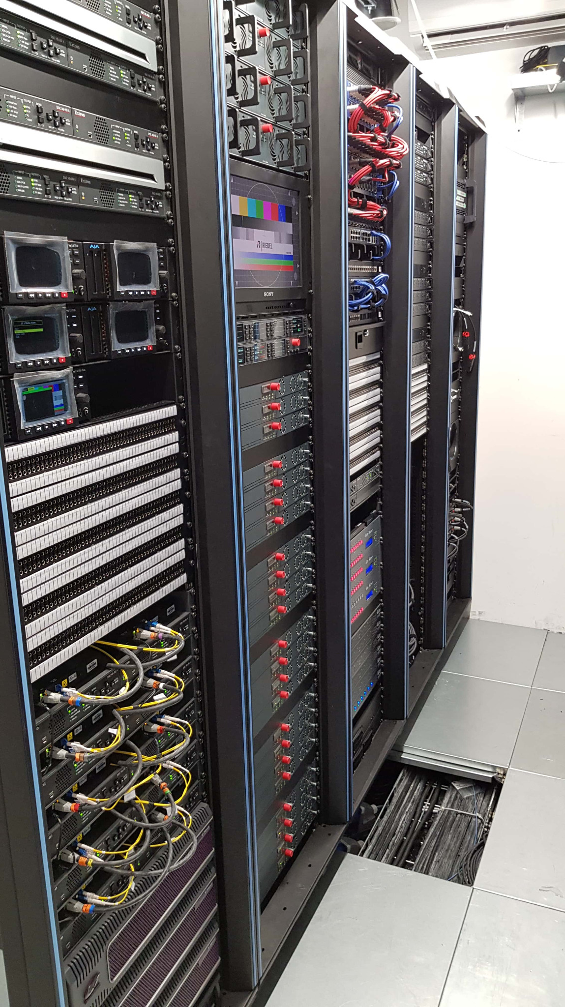 Rząd szaf serwerowych z widocznymi kablami i urządzeniami sieciowymi, w tle ekran Sony z kolorowymi paskami testowymi Riedel, pod podłogą techniczną widoczna wiązka kabli.