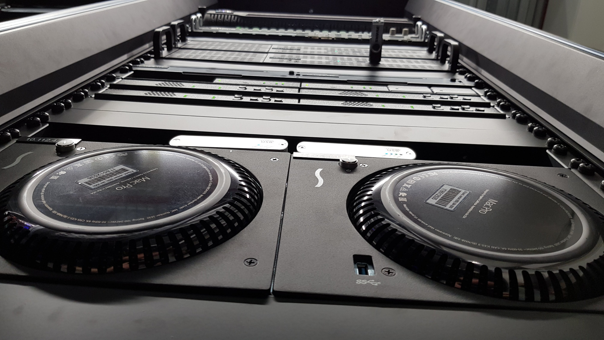 Dwa komputery Mac Pro umieszczone w serwerowni, widoczne w perspektywie od dołu, z widocznym rackiem i innymi urządzeniami serwerowymi w tle.