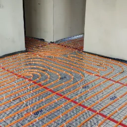 Pomarańczowe rury grzewcze podłogowe rozłożone na folii z siatką w surowym wnętrzu budynku w trakcie instalacji.