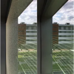 Porównanie okna przed i po czyszczeniu, widoczne różnice w przejrzystości szyby i czystości ramy okiennej, z widokiem na budynek biurowy i parking.