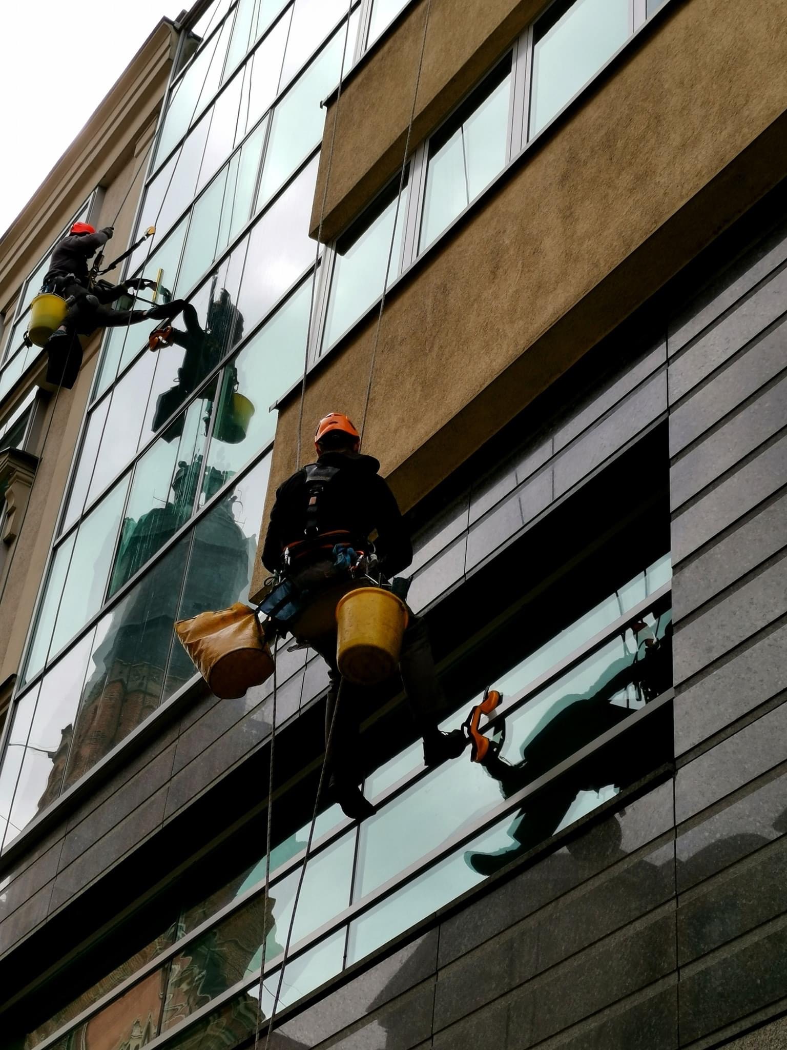Dwóch alpinistów przemysłowych myje okna biurowca, widoczne odbicie budynku w oknach.
