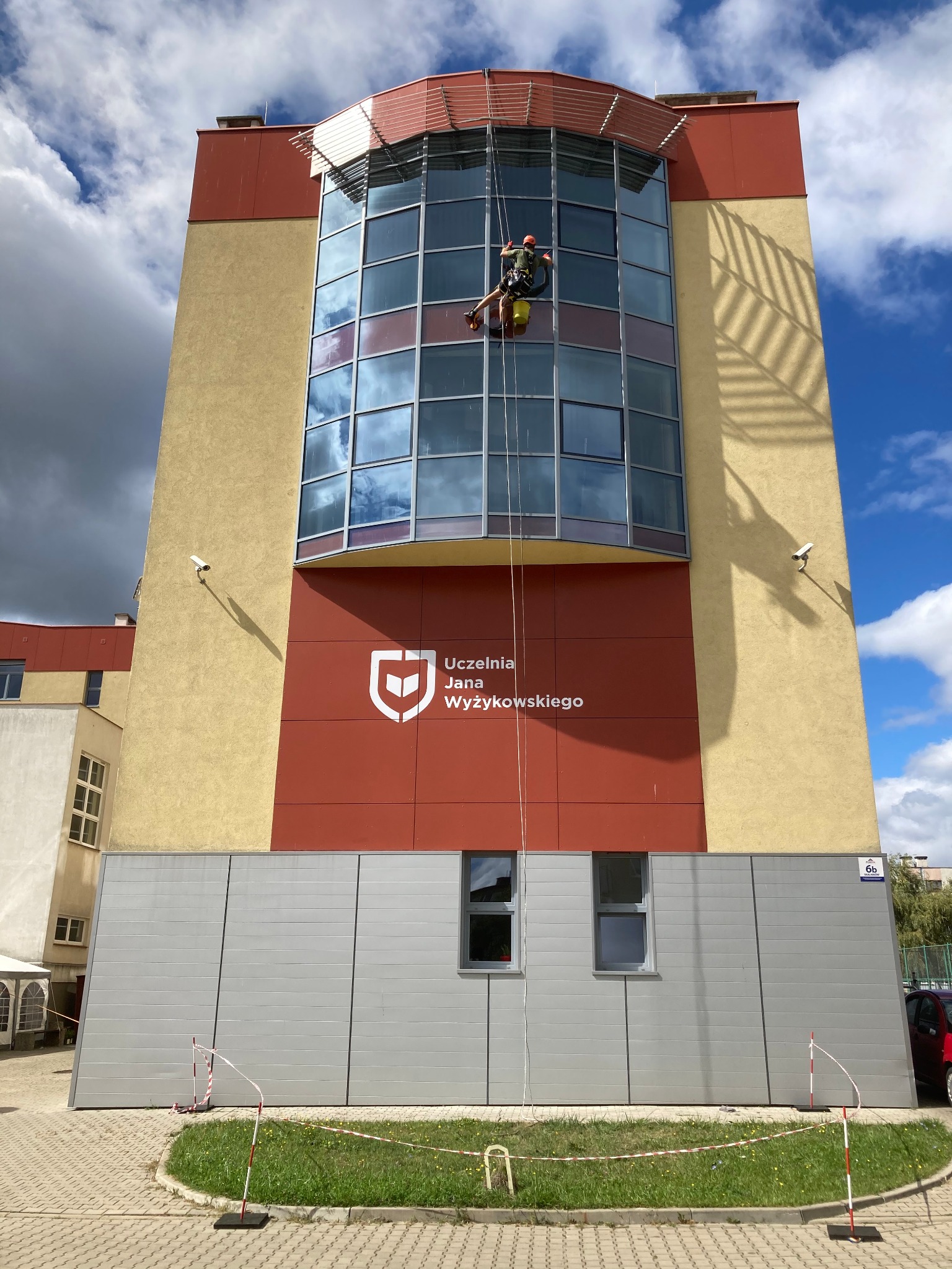 Pracownik na linach zabezpieczających czyści okna na wysokości budynku Uczelni Jana Wyżykowskiego. Widoczne odbicia chmur w oknach i żółte wiadro.