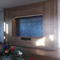 Duża, drewniana zabudowa meblowa w salonie z miejscem na telewizor, oświetleniem LED i szarą ścianą w tle, dekoracja świąteczna w pierwszym planie.