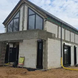 Dom w trakcie budowy z zamontowanymi oknami i roletami zewnętrznymi w czarnych ramach, widoczne rusztowania i materiały budowlane, żółte rury wentylacyjne leżące na piasku.