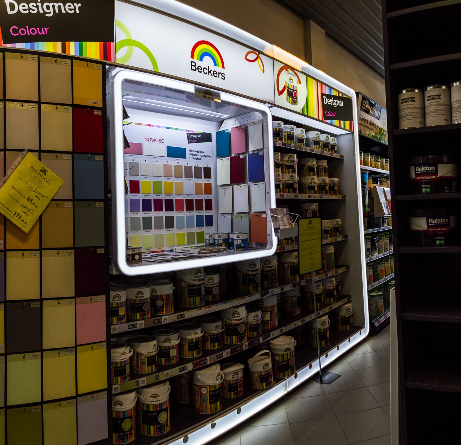 Ekspozycja farb marki Beckers w sklepie budowlanym, widoczne puszki z farbami Designer Colour na półkach z podświetleniem LED oraz wzorniki kolorów na ścianie.