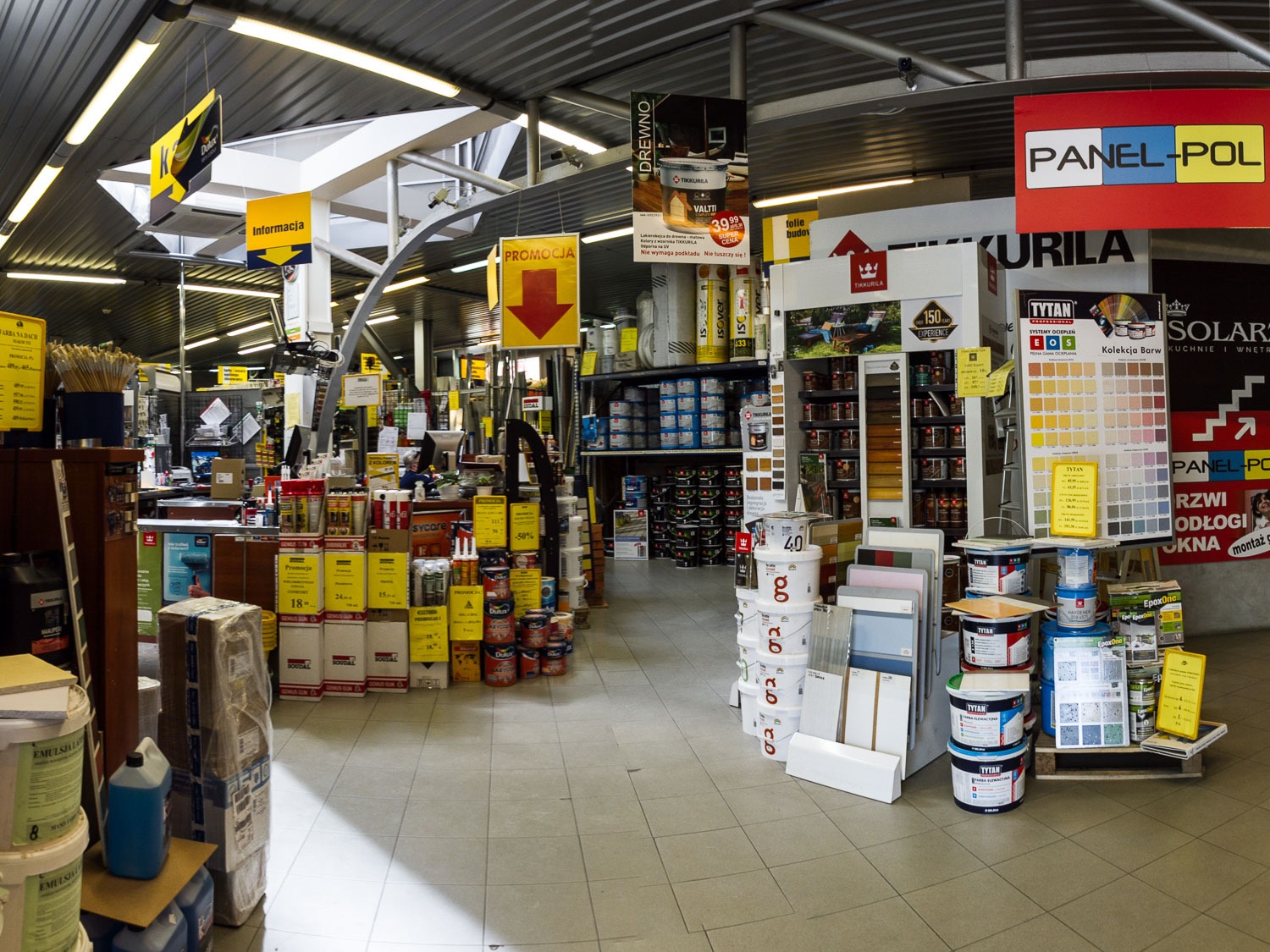 Wnętrze hali z materiałami budowlanymi, regały wypełnione farbami, lakierami, chemią budowlaną, widoczne oznaczenia promocyjne i informacyjne, oświetlenie sufitowe.
