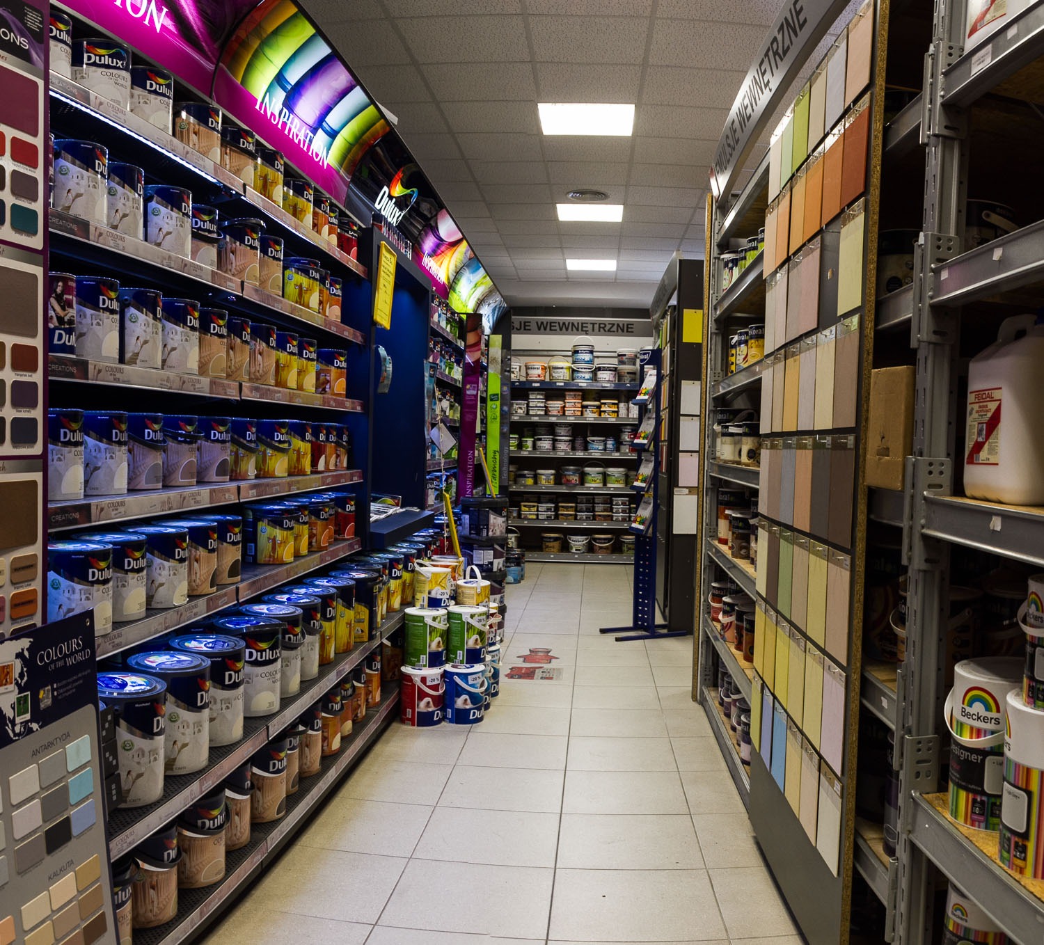 Wnętrze sklepu z farbami: regały pełne puszek farb Dulux i Beckers, wzorniki kolorów na ścianach, oświetlenie sufitowe, widok w głąb alejki między regałami.