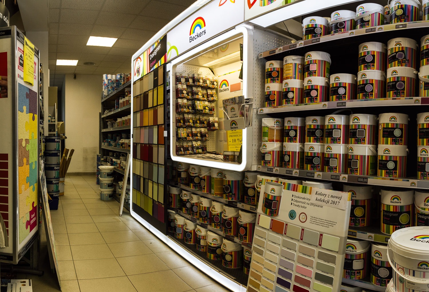 Półki w sklepie z farbami Beckers, prezentujące szeroką gamę kolorów i odcieni w puszkach, wraz z ekspozytorem próbek kolorystycznych i tabliczką informacyjną o promocji.