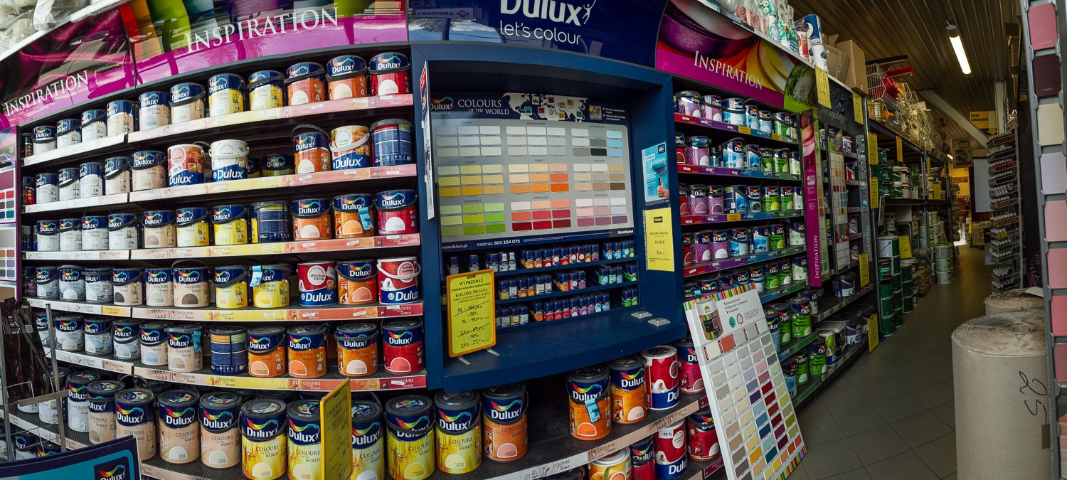 Wnętrze sklepu w Krakowie z półkami pełnymi puszek farb Dulux w różnych kolorach. Widoczny ekspozytor z próbkami kolorów i promocją cenową.