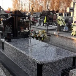 Granitowy nagrobek z czarnymi akcentami na cmentarzu, w tle drzewa o jesiennych barwach i inne groby.