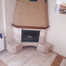 Kącik z marmurowym kominkiem o geometrycznej bryle, wykończony drewnianym gzymsem, dopasowany kolorystycznie do podłogi z płytek imitujących marmur.