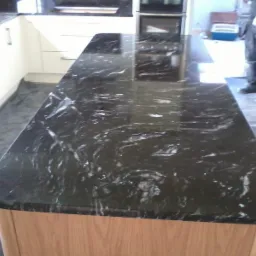 Czarny, polerowany blat z kamienia naturalnego z wyraźnym, białym żyłkowaniem, zamontowany na drewnianej wyspie kuchennej; w tle widoczna zabudowa kuchenna i pracownik w kombinezonie.