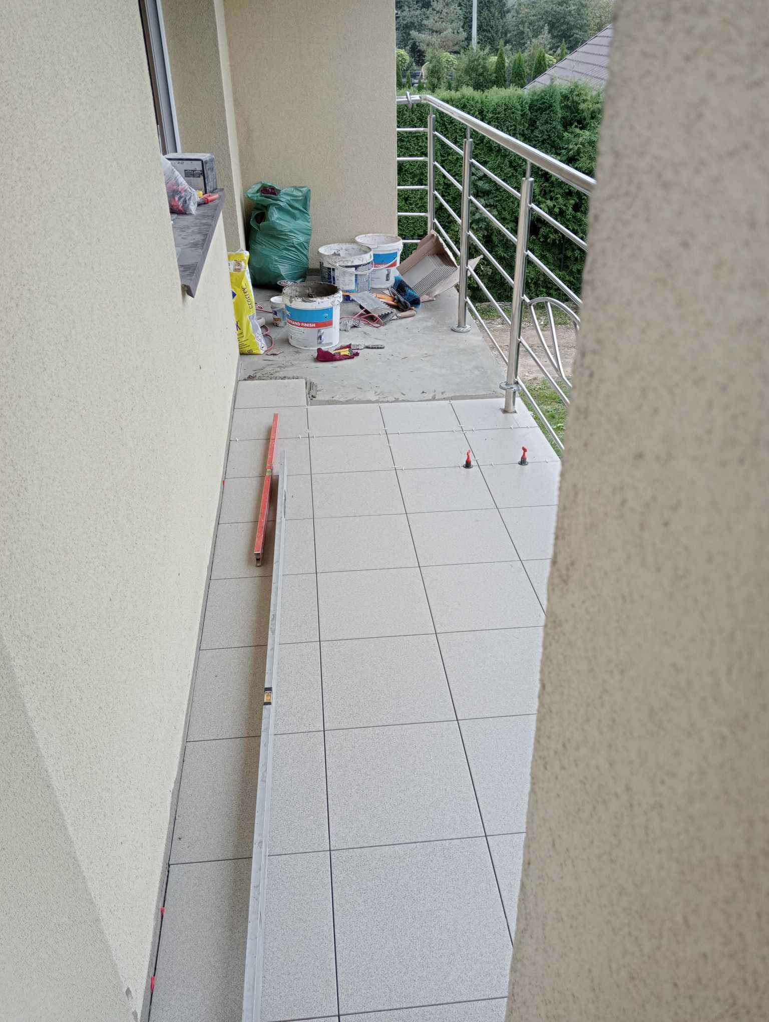 Balkon w trakcie kładzenia jasnych, kwadratowych płytek ceramicznych, widoczne poziomice, wiaderka po zaprawie i torba z klejem, balustrada ze stali nierdzewnej.