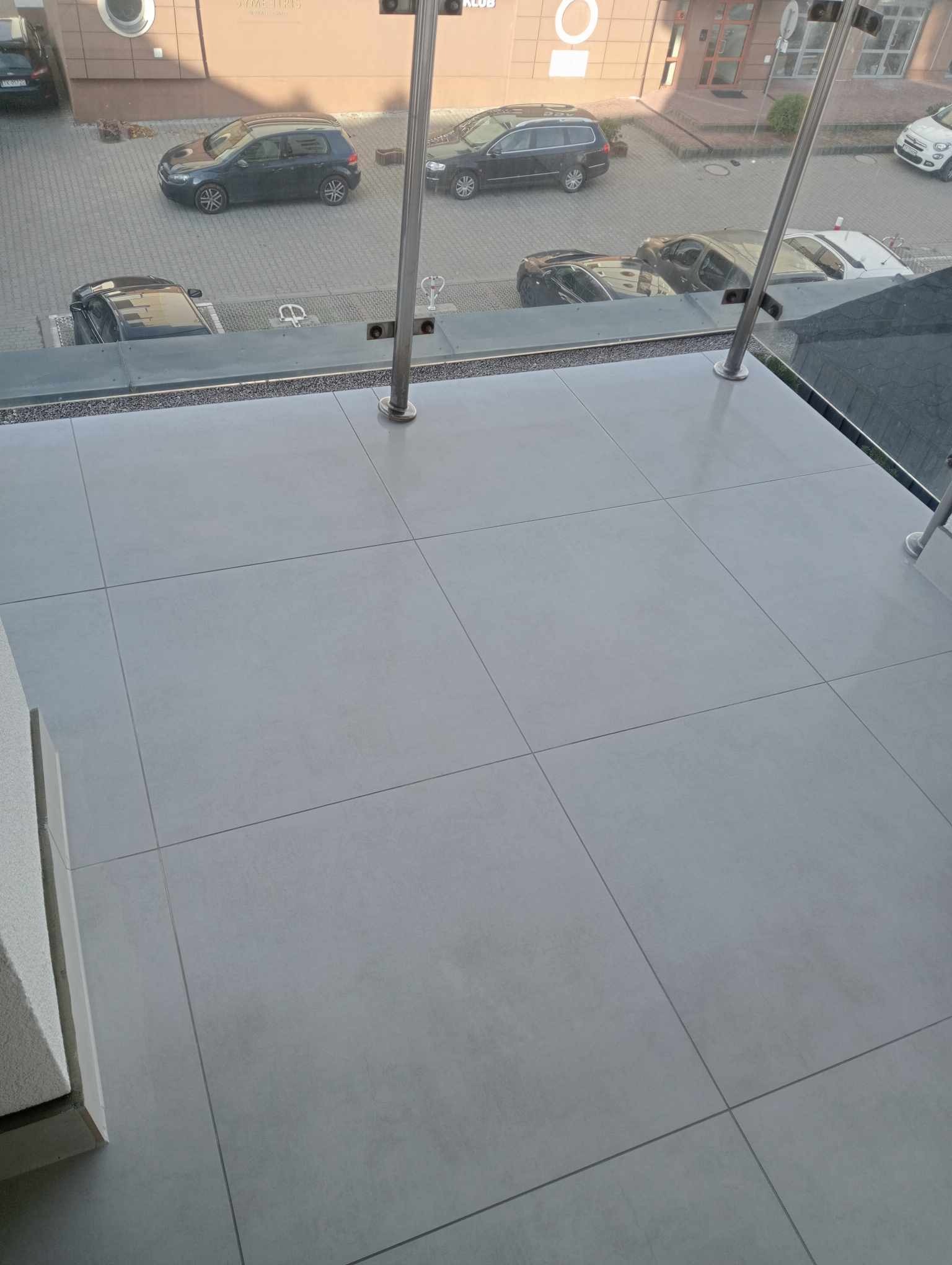 Jasnoszare płytki podłogowe na balkonie z widokiem na parking i budynek z przeszkloną balustradą.