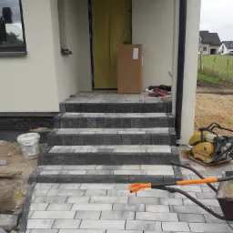 Schody wejściowe do domu wyłożone szarą kostką brukową, widoczna ubijarka wibracyjna, taczka z piaskiem i łopata z pomarańczowym uchwytem.