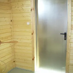 Wnętrze kabiny sanitarnej z drewnianymi ścianami, metalowymi drzwiami z czarną klamką i drewnianym wieszakiem na ręczniki.