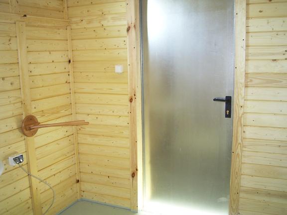 Wnętrze kabiny sanitarnej z drewnianymi ścianami, metalowymi drzwiami z czarną klamką i drewnianym wieszakiem na ręczniki.