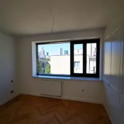 Pusty pokój z parkietem w jodełkę, widokiem na panoramę miasta przez okno i białą szafą w zabudowie.