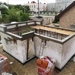 Wznoszenie ścian parterowego budynku z betonu komórkowego, widoczne zbrojenia i deskowanie stropu, paleta z cegłami klinkierowymi na pierwszym planie.