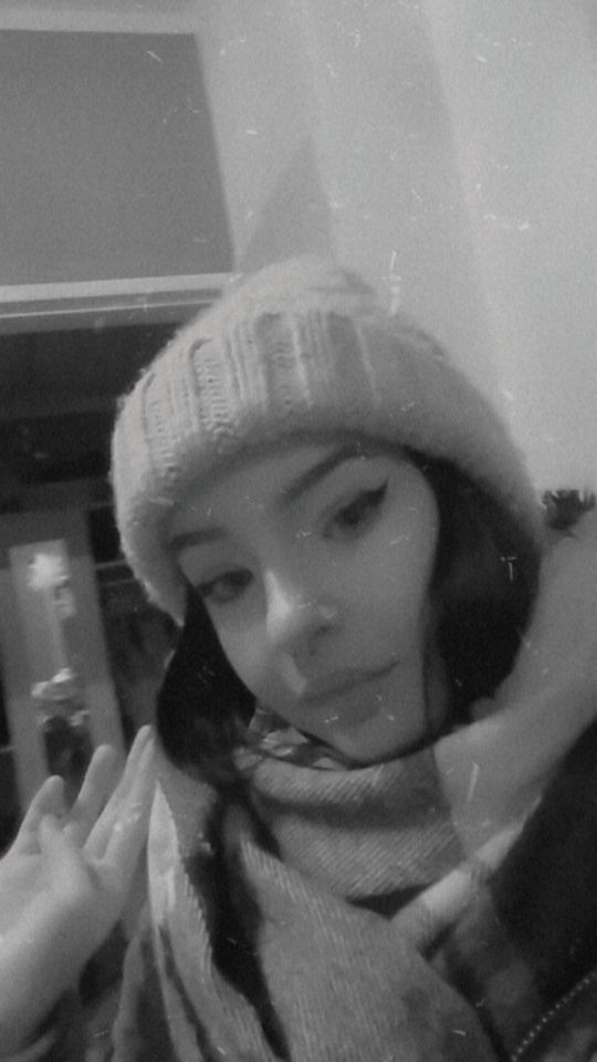 Czarno-białe selfie kobiety w czapce i szaliku, z mocnym makijażem oczu, w słabym oświetleniu.