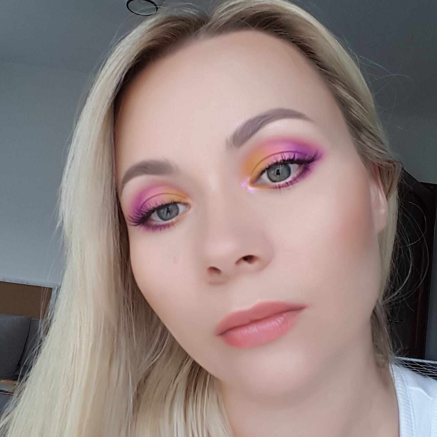 Portret blondwłosej kobiety z profesjonalnie wykonanym, kolorowym make-upem oczu w odcieniach różu, fioletu i żółci, wykonanym w Wieliczce, z widocznymi sztucznymi rzęsami i delikatnym makijażem ust.