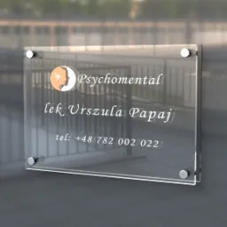 Elegancka, przezroczysta tabliczka informacyjna z logo i danymi kontaktowymi firmy 'Psychomental' zamocowana na zewnątrz budynku.