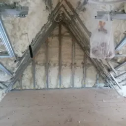 Poddasze z ociepleniem pianą PUR, widoczne metalowe profile konstrukcyjne i fragment drewnianej podłogi, z workiem foliowym wiszącym na jednym z profili.